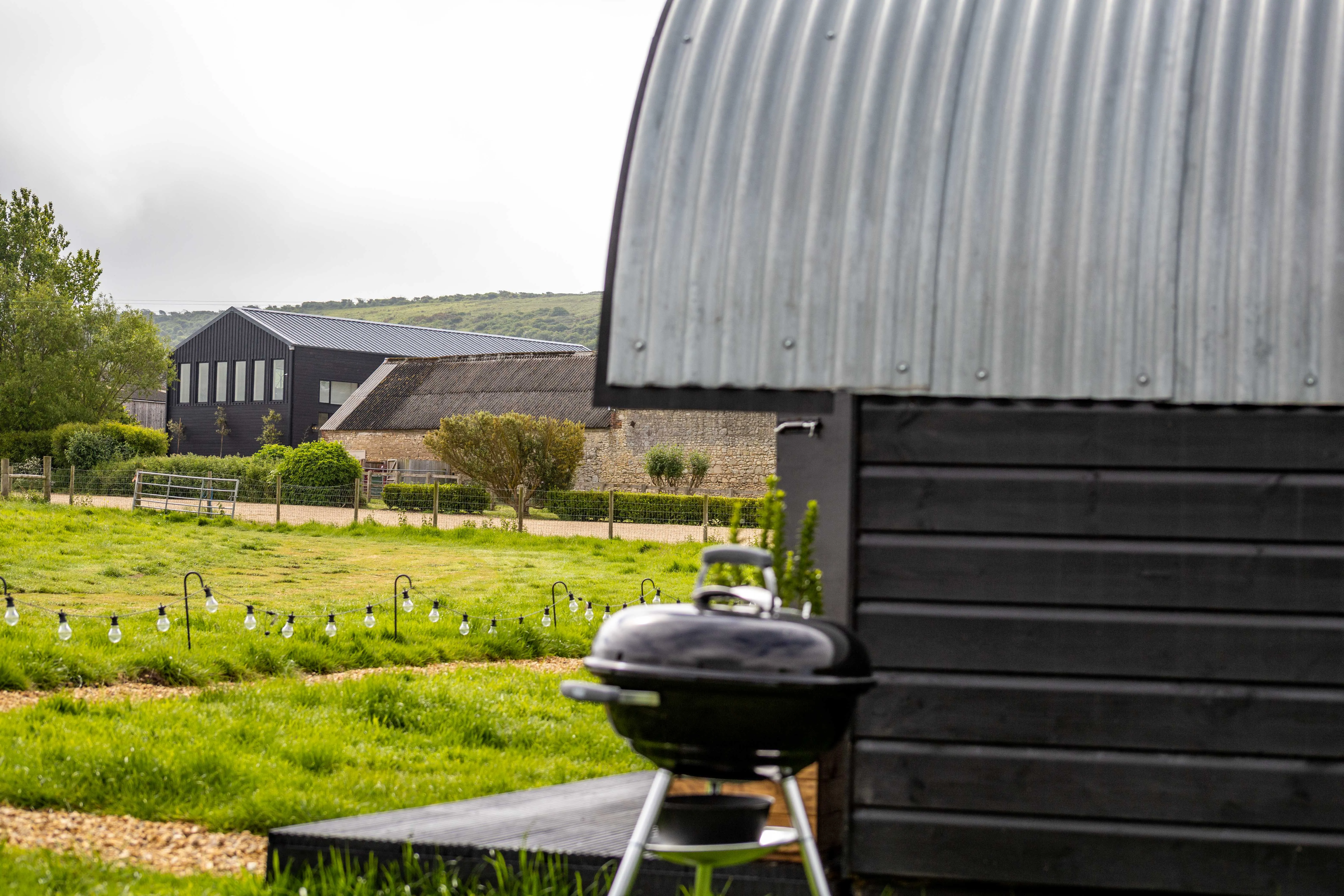 Countryside view with a black timber hut in the foreground, a barbecue on the grass, and farm buildings in the distance