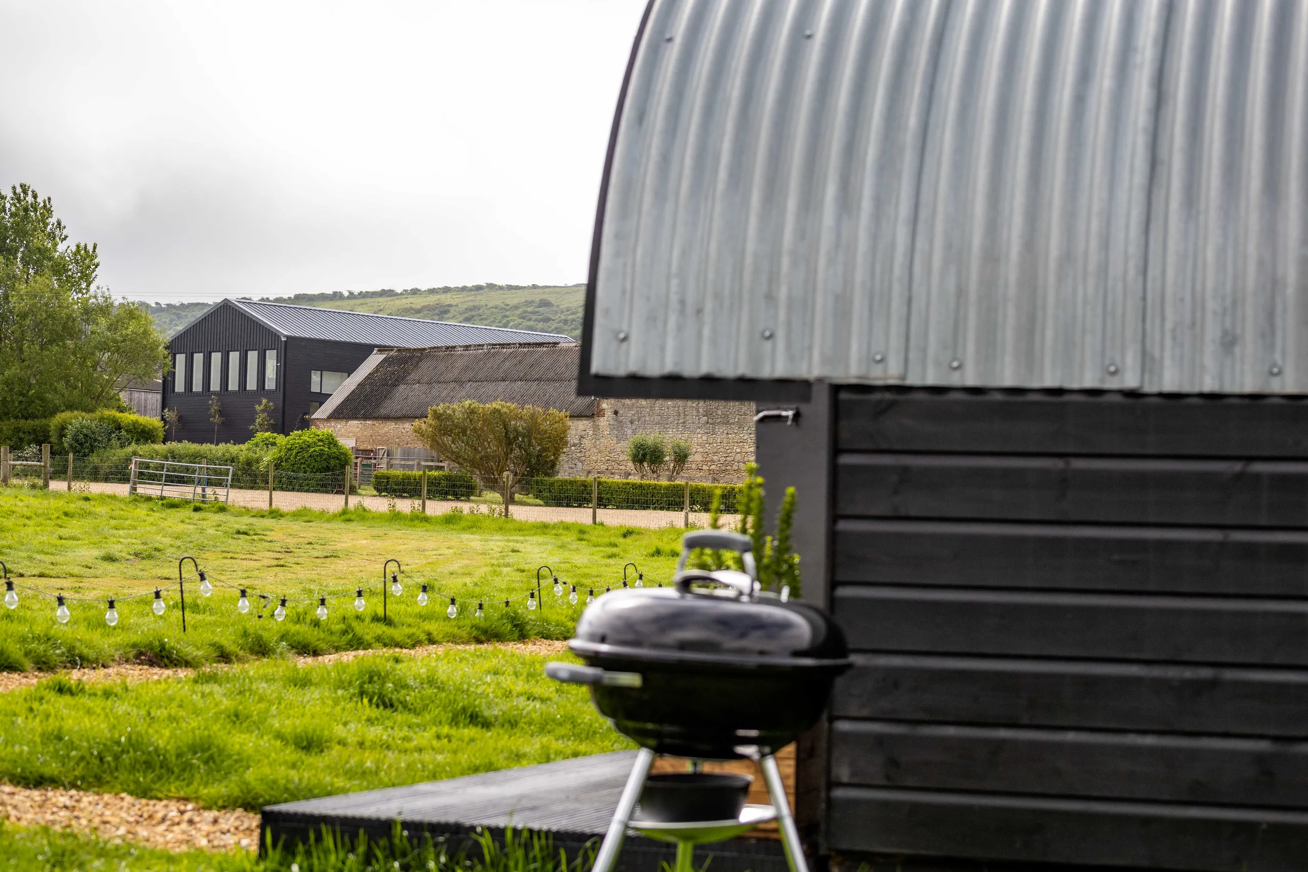 Countryside view with a black timber hut in the foreground, a barbecue on the grass, and farm buildings in the distance
