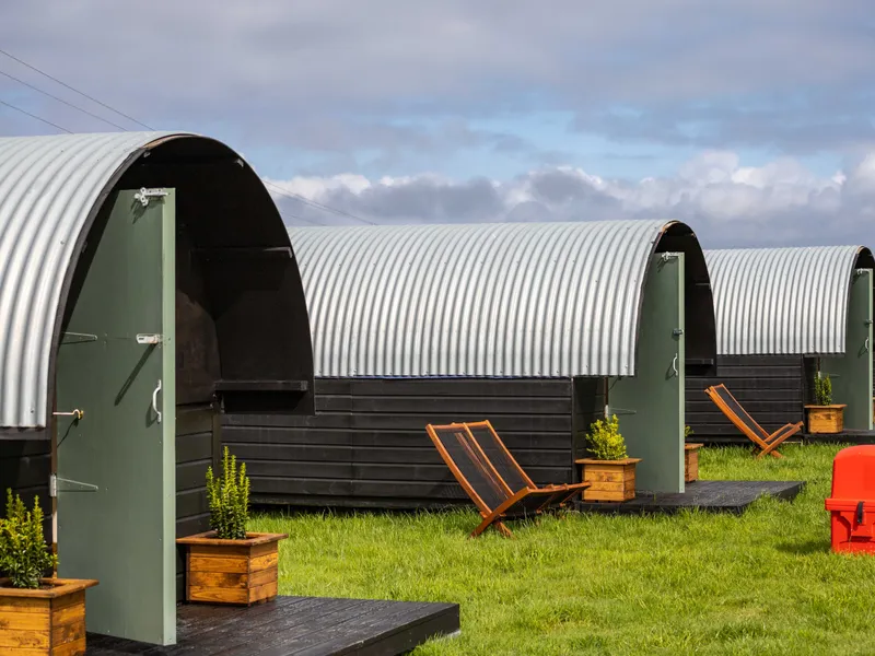 Row of corrugated metal hut-style cabins with green doors and deckchairs on a grassy field