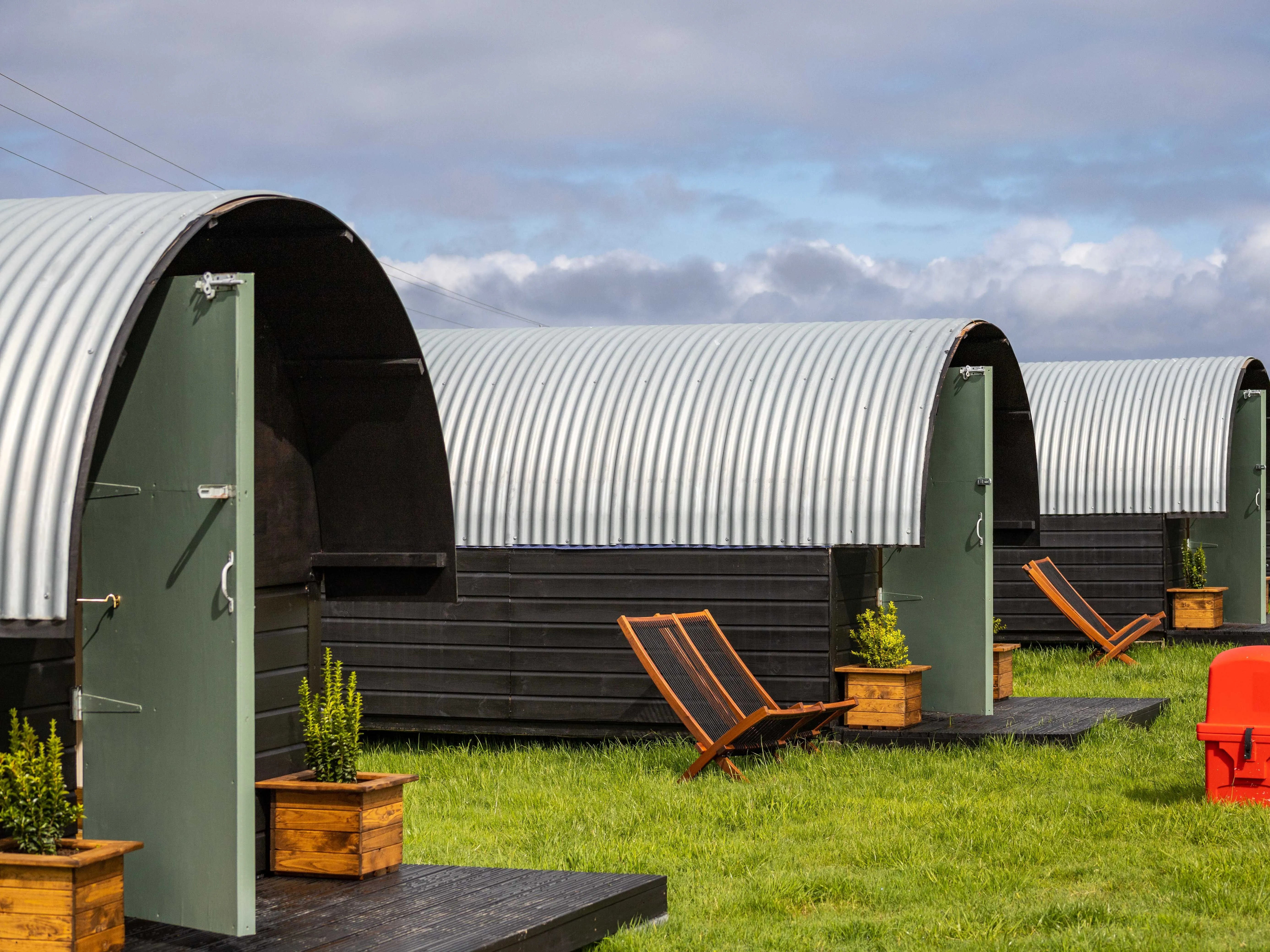 Row of corrugated metal hut-style cabins with green doors and deckchairs on a grassy field