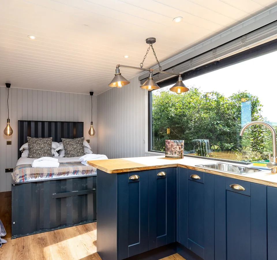 Cosy shepherd’s hut interior with double bed and compact kitchen, blue cabinetry and large window overlooking greenery