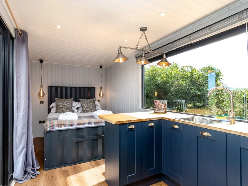 Cosy shepherd’s hut interior with double bed and compact kitchen, blue cabinetry and large window overlooking greenery