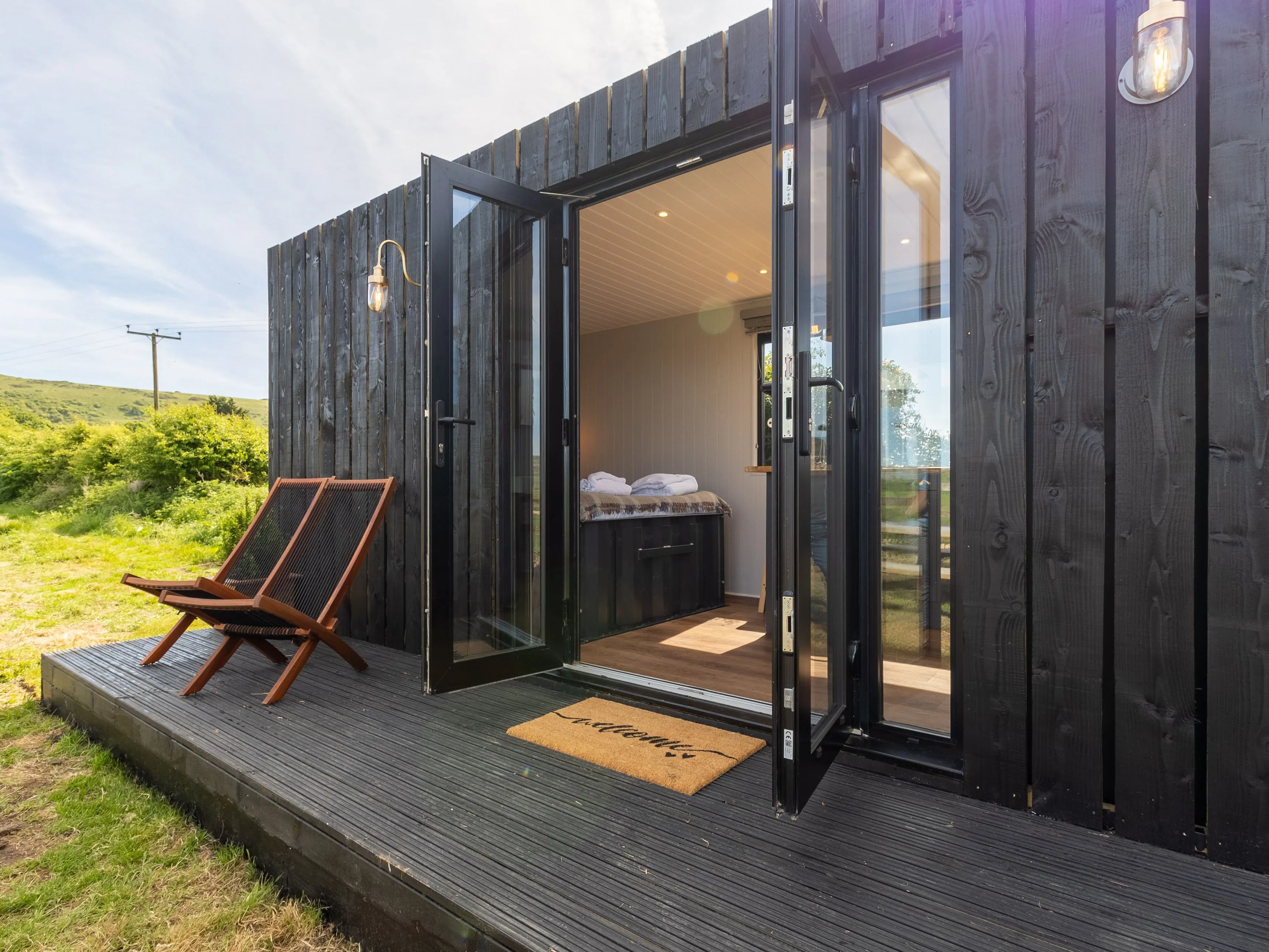 Black timber cabin with open glass doors onto a small deck with two loungers, set in green countryside