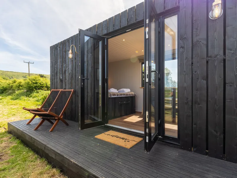 Black timber cabin with open glass doors onto a small deck with two loungers, set in green countryside