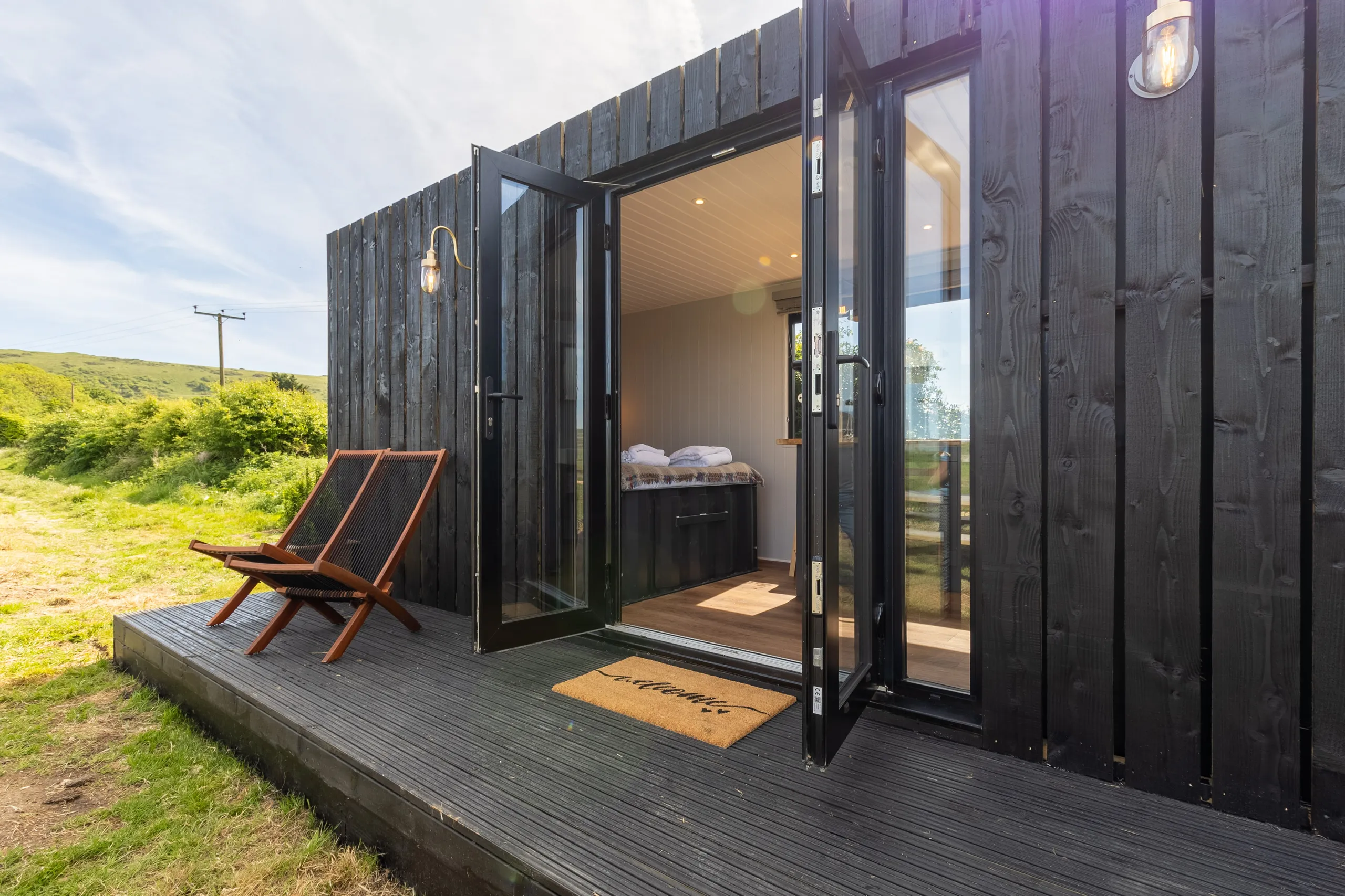 Black timber cabin with open glass doors onto a small deck with two loungers, set in green countryside