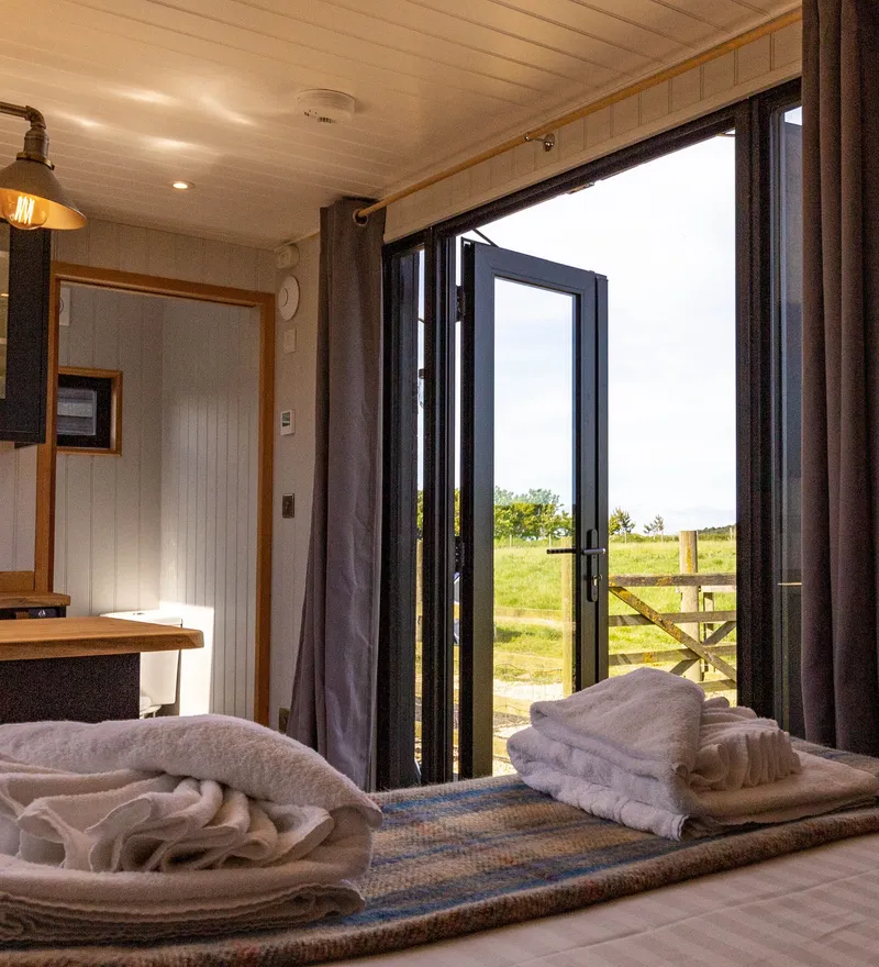 Cosy cabin bedroom with folded towels on the bed and open glass doors to countryside views