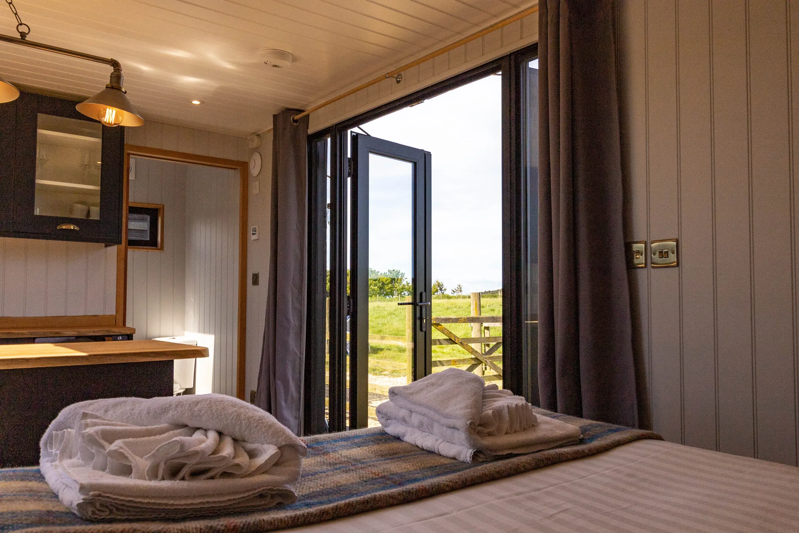 Cosy cabin bedroom with folded towels on the bed and open glass doors to countryside views