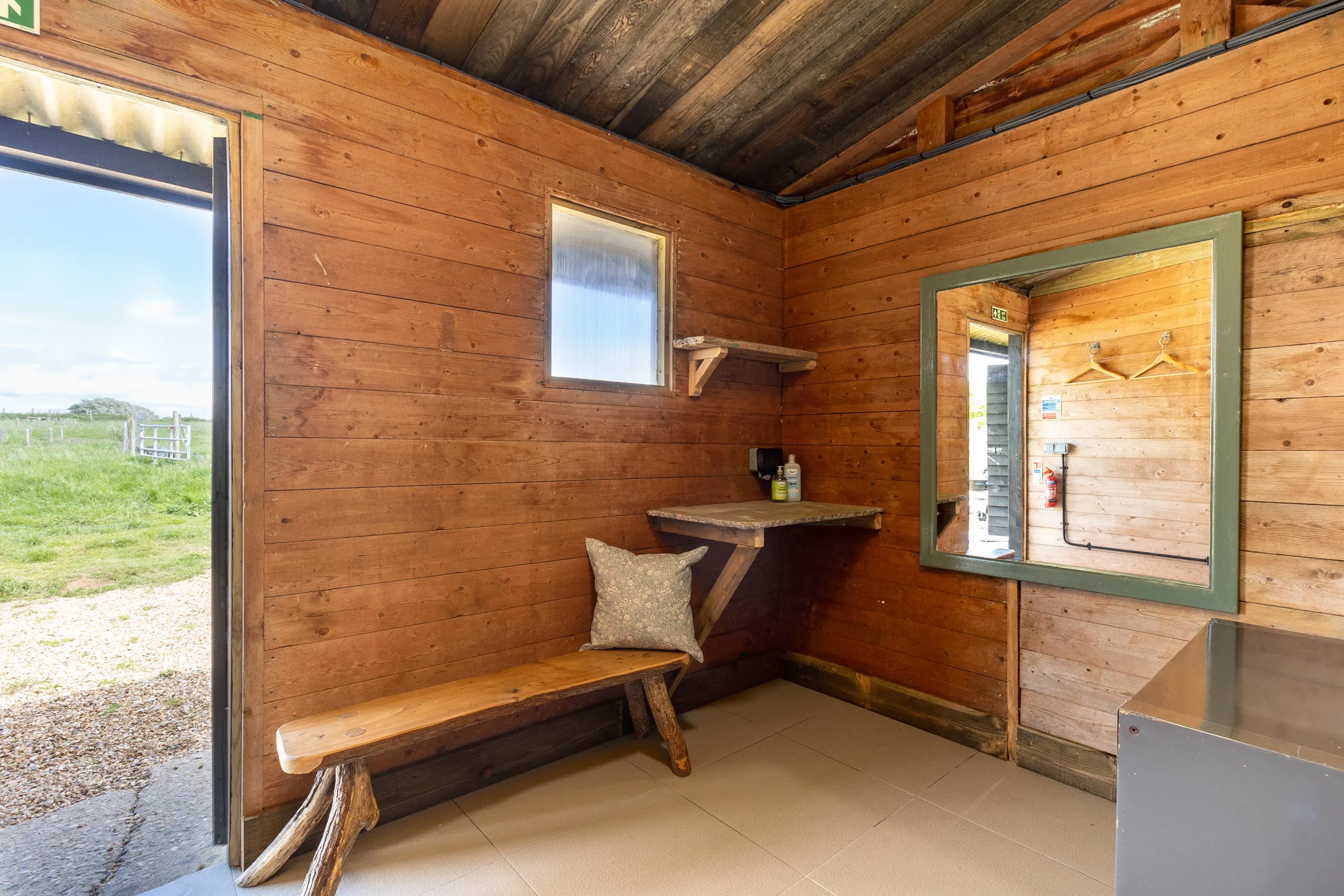 Wood-panelled cabin interior with rustic bench, small shelf and mirror by an open doorway to a grassy field