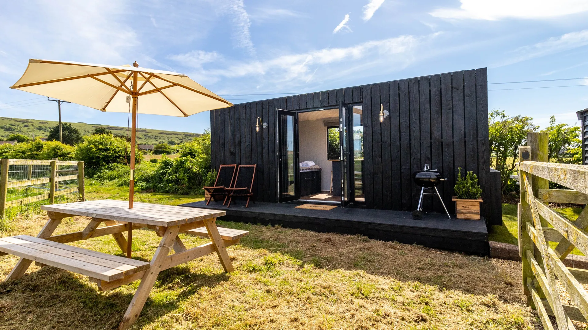 A small black wooden cabin with open French doors, a picnic table and parasol in a sunny countryside garden.