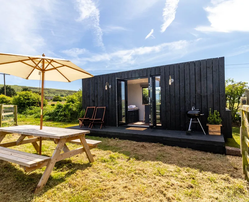 A small black wooden cabin with open French doors, a picnic table and parasol in a sunny countryside garden.