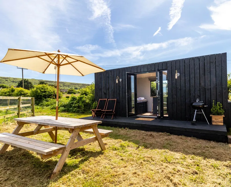 A small black wooden cabin with open French doors, a picnic table and parasol in a sunny countryside garden.