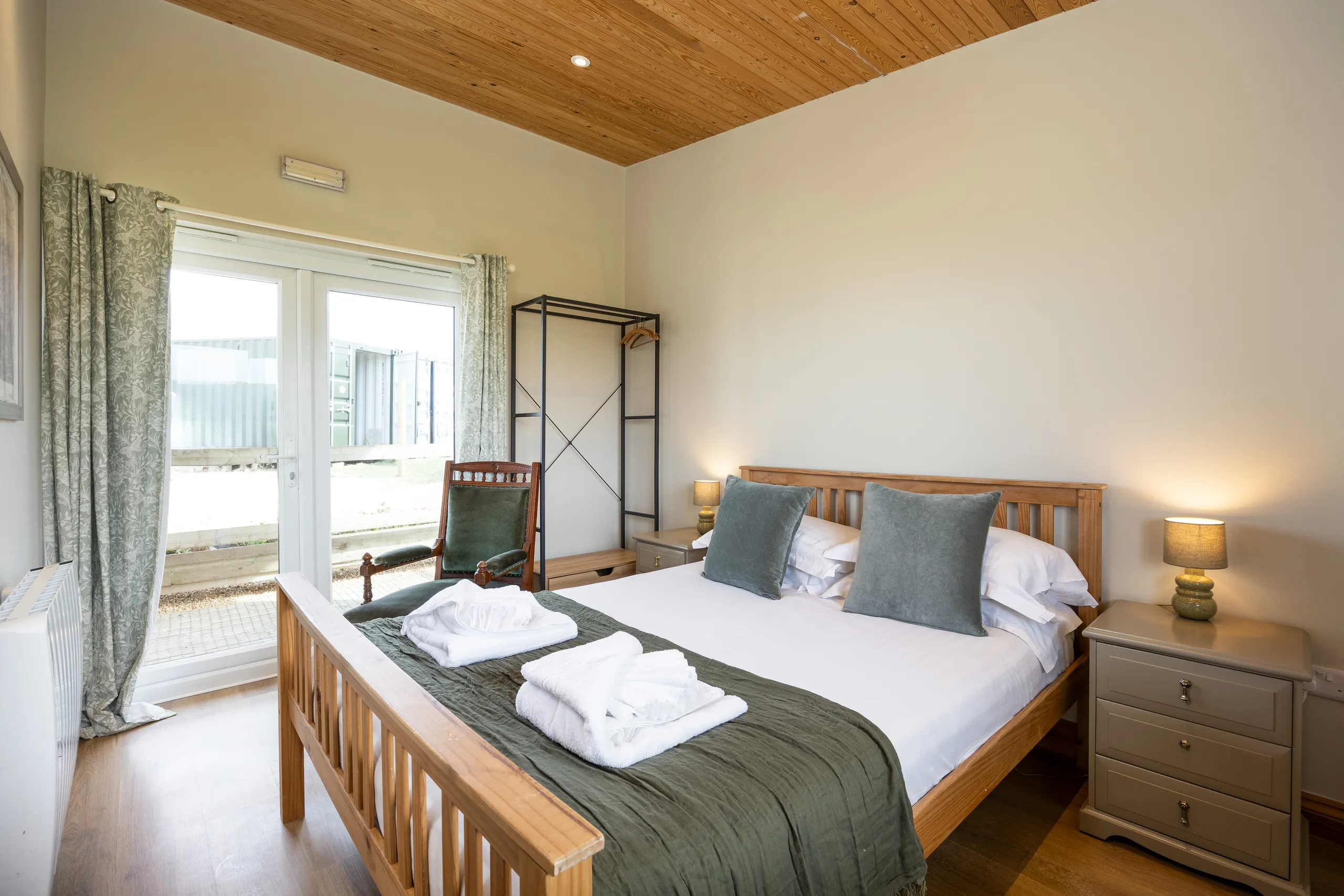 Light and airy bedroom with wooden bed, green cushions, bedside lamps and French doors with patterned curtains