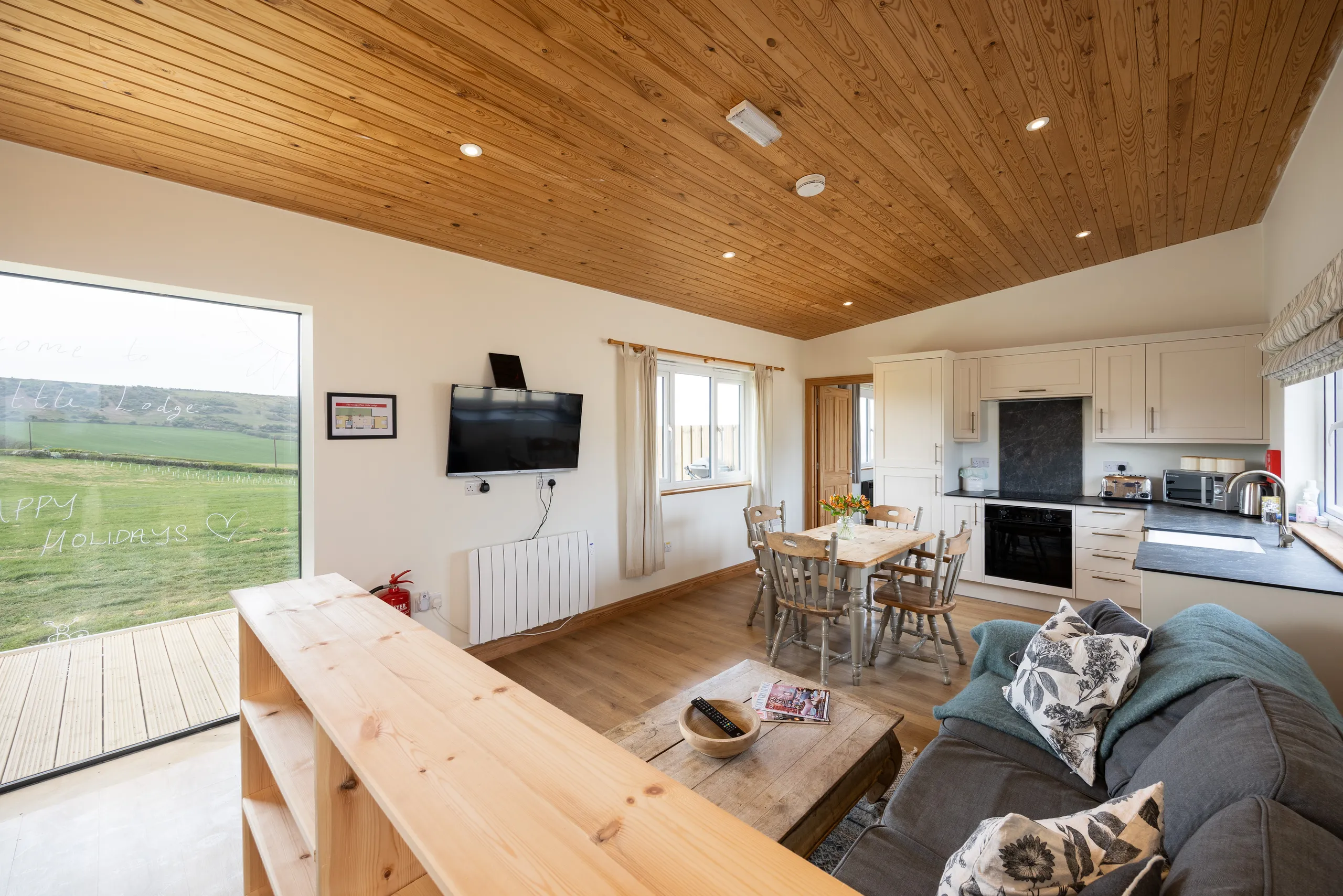 Open-plan lodge living area with kitchen, dining table, sofa and countryside view through sliding doors