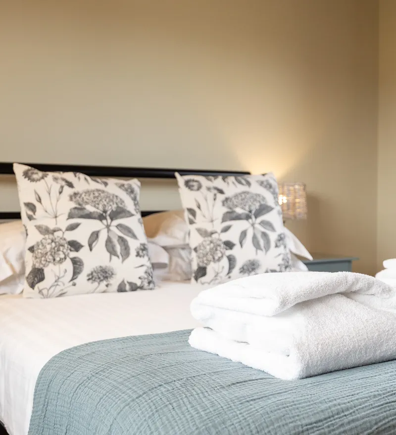 Cosy bedroom with double bed, floral cushions and folded white towels on a blue throw