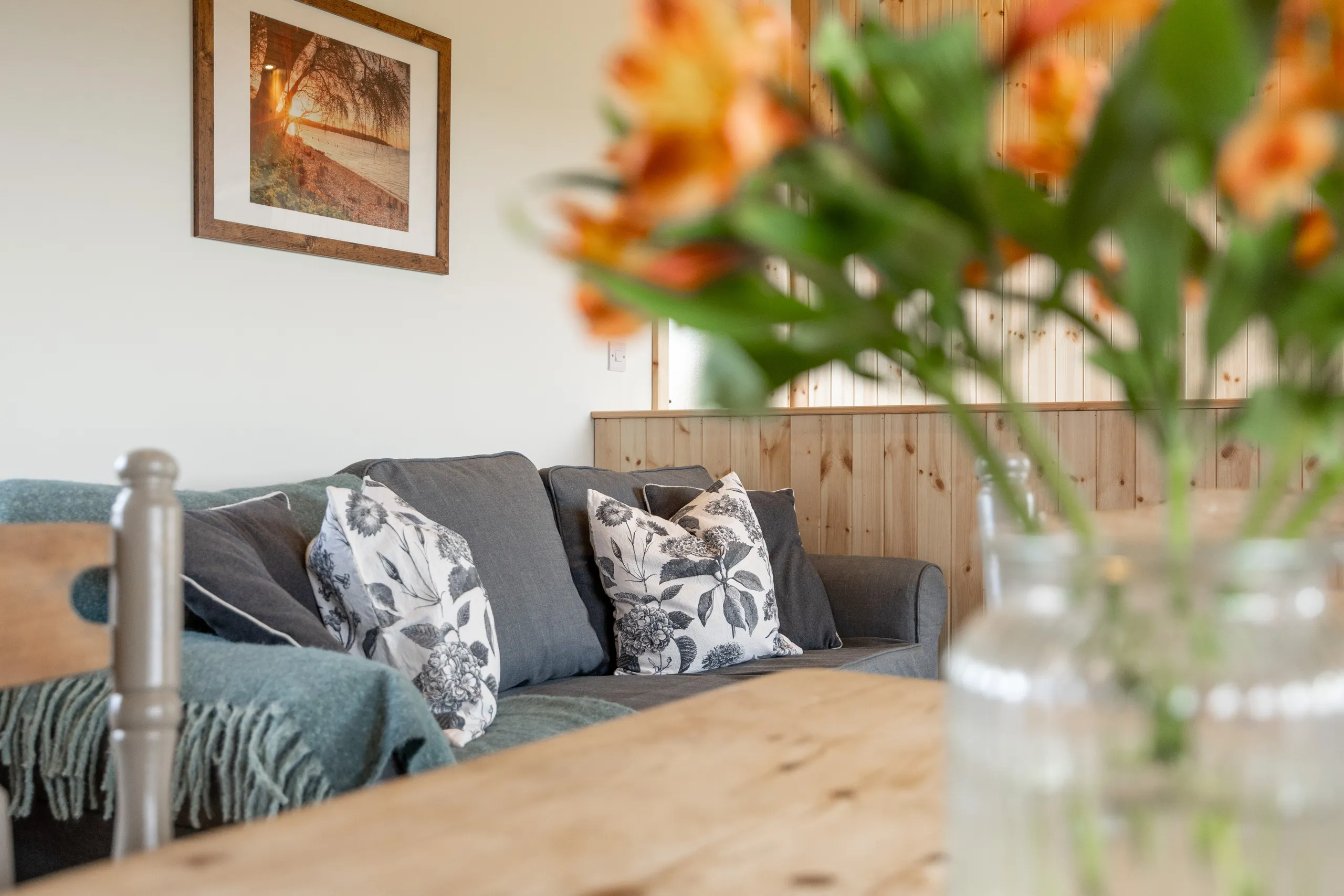 Cosy lodge living area with grey sofa, floral cushions, wood-panelled wall and a blurred vase of orange flowers in the foreground