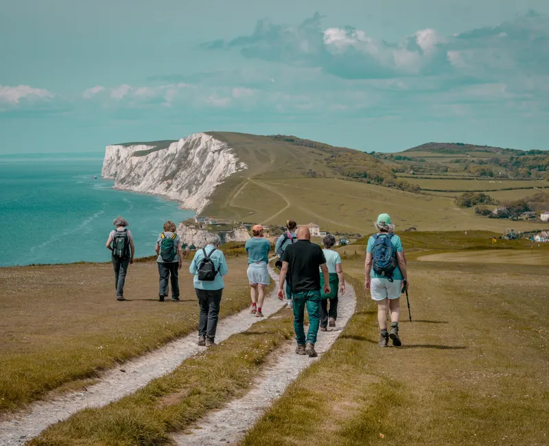 Group of hikers walking along a coastal path towards white chalk cliffs beside the sea