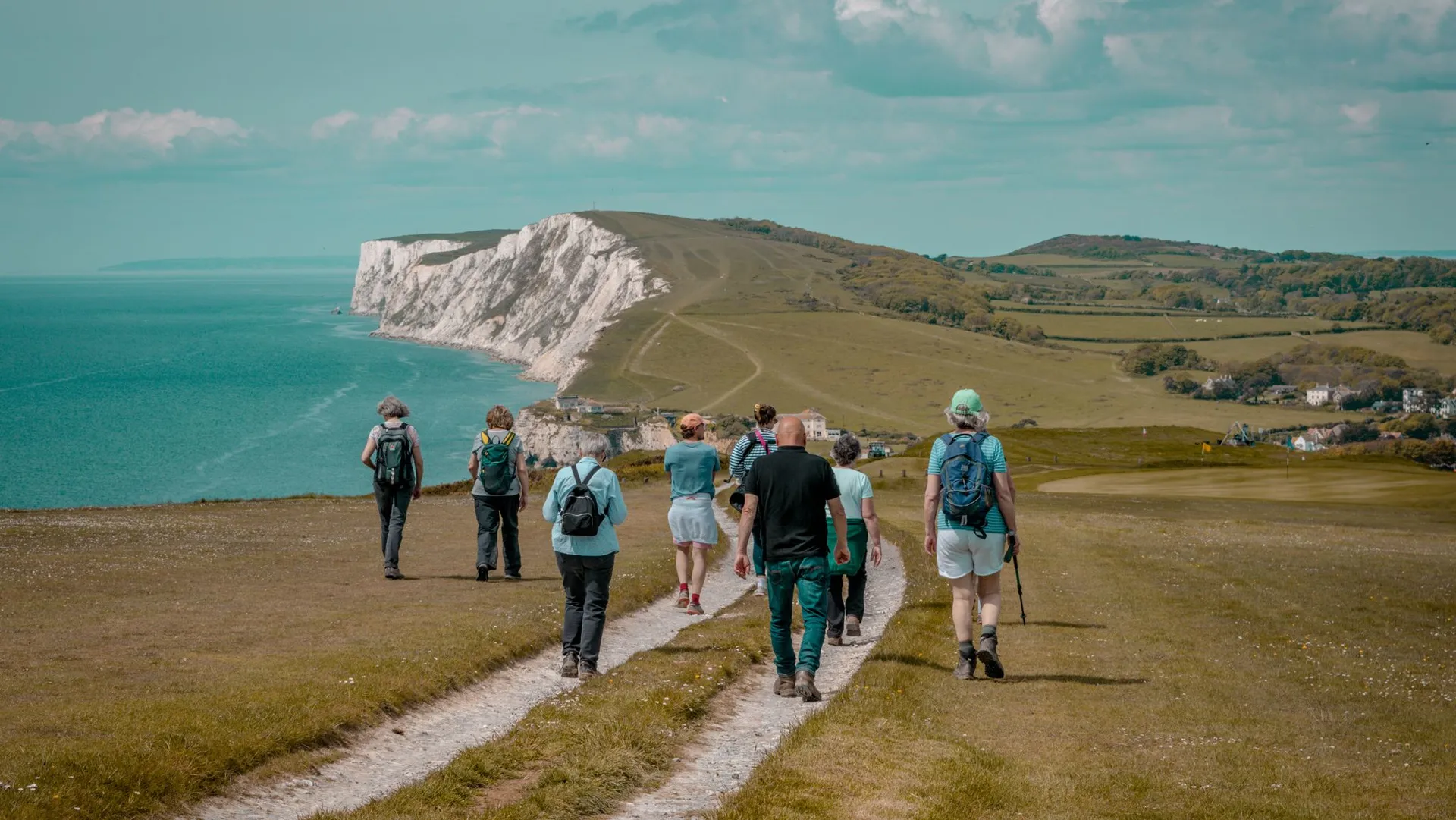 Group of hikers walking along a coastal path towards white chalk cliffs beside the sea