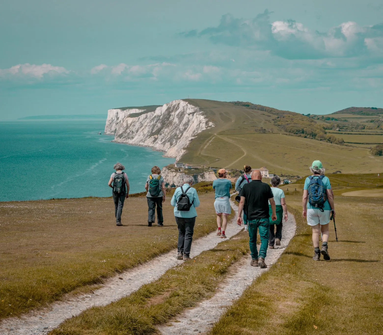 Group of hikers walking along a coastal path towards white chalk cliffs beside the sea