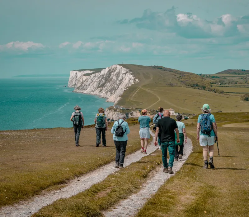 Group of hikers walking along a coastal path towards white chalk cliffs beside the sea