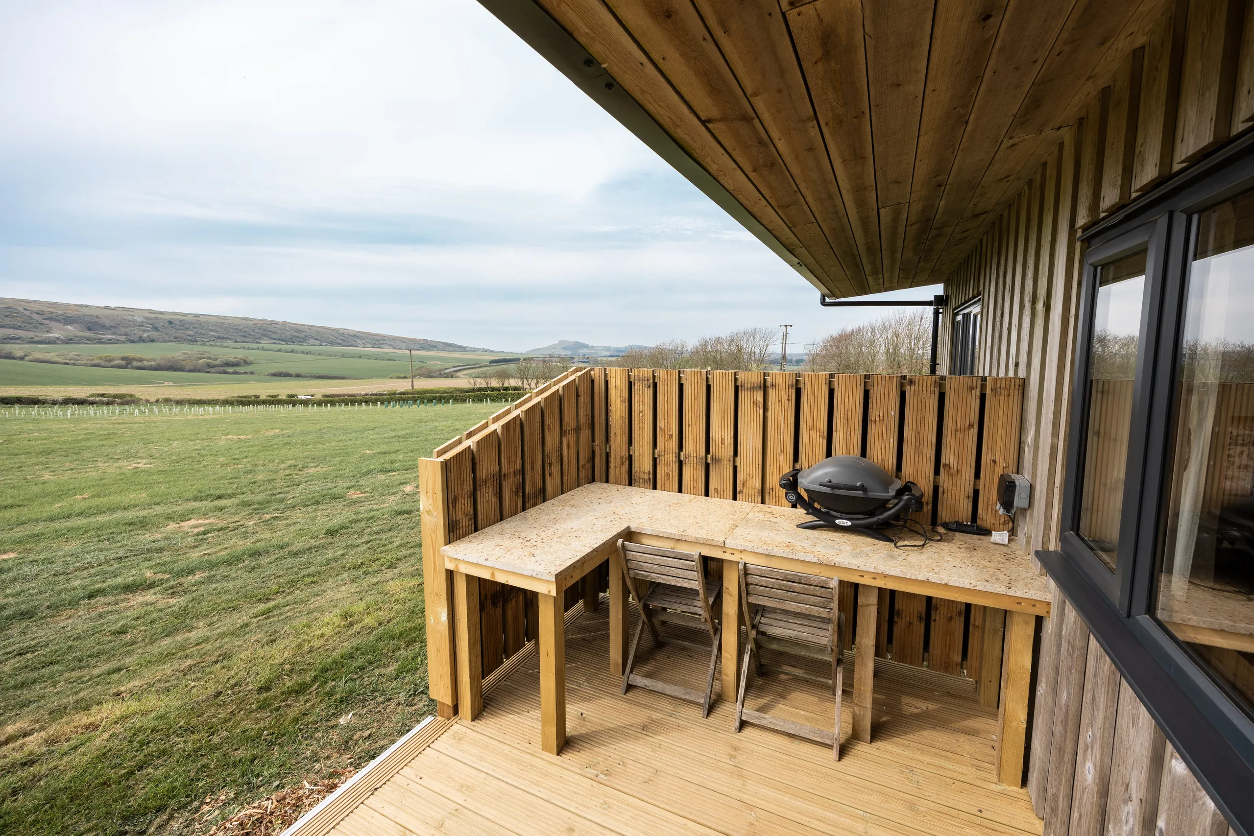 Outdoor deck with wooden table, two chairs and a barbecue overlooking rolling countryside