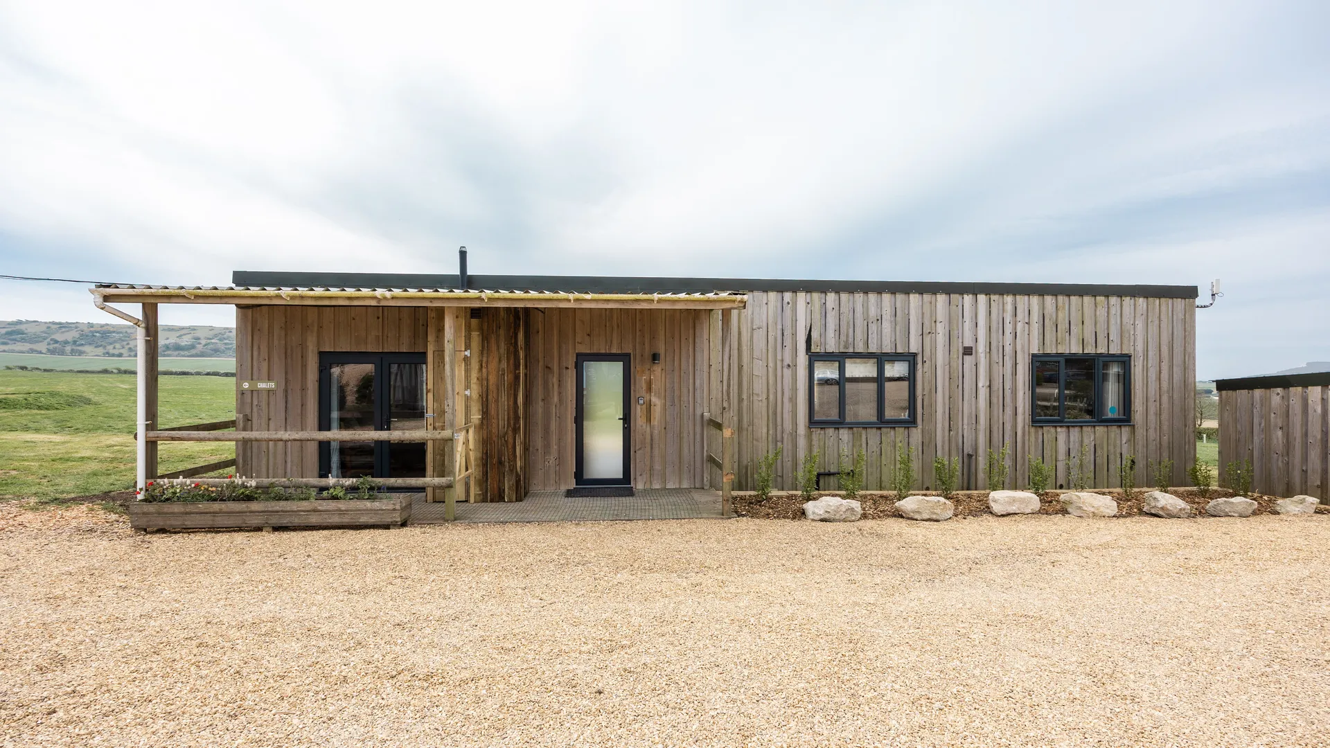 Exterior of a modern timber-clad lodge with a covered porch, set on a gravel area overlooking green countryside.