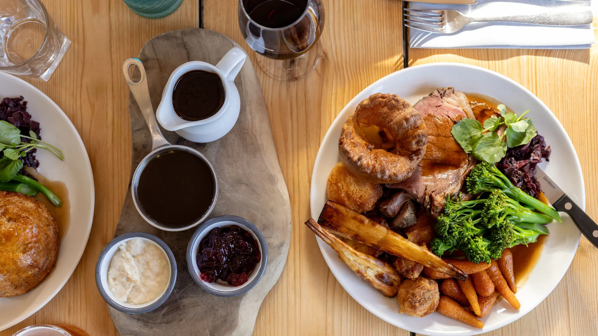Overhead view of a roast dinner with Yorkshire pudding, roast potatoes, carrots, greens and gravy on a wooden table.