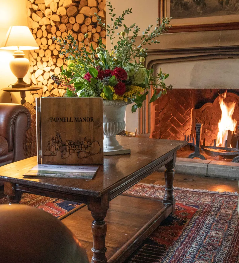 Cosy lounge with leather sofa, wooden coffee table, floral arrangement and a lit fireplace at Tapnell Manor
