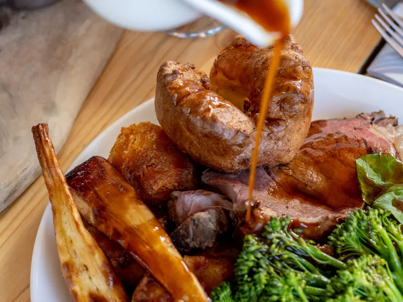 Gravy being poured over a roast dinner with Yorkshire pudding, roast potatoes, carrots and broccoli