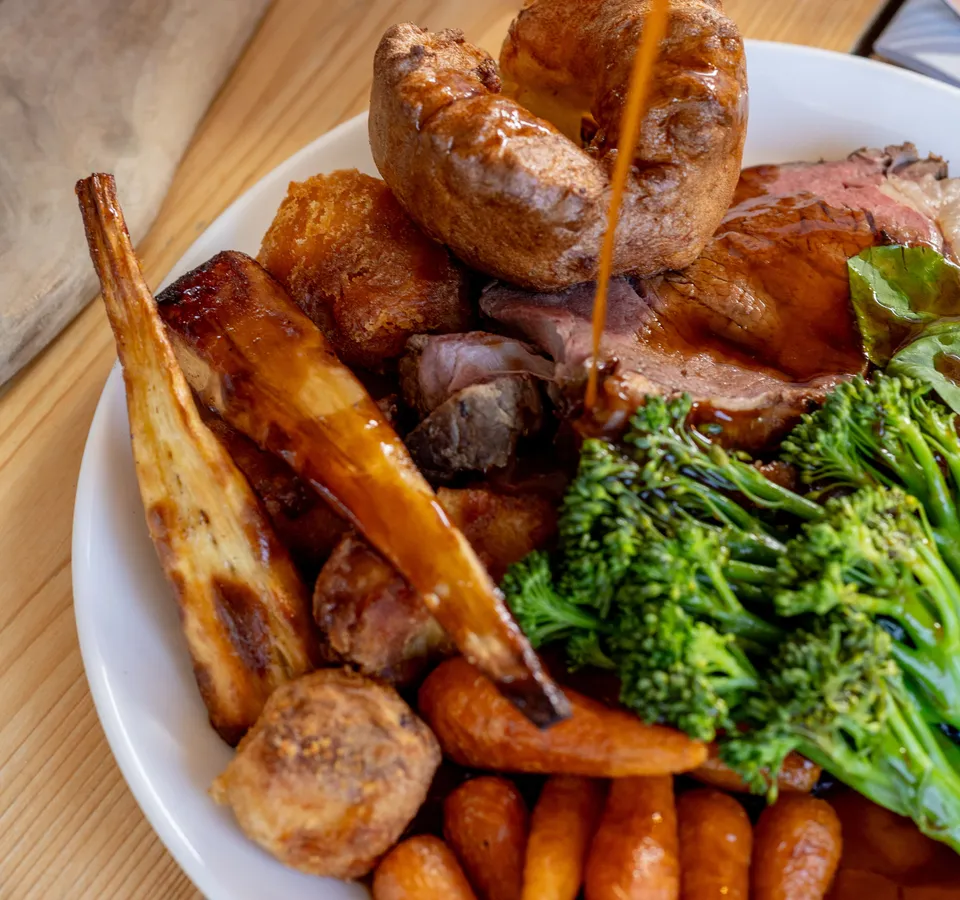 Gravy being poured over a roast dinner with Yorkshire pudding, roast potatoes, carrots and broccoli