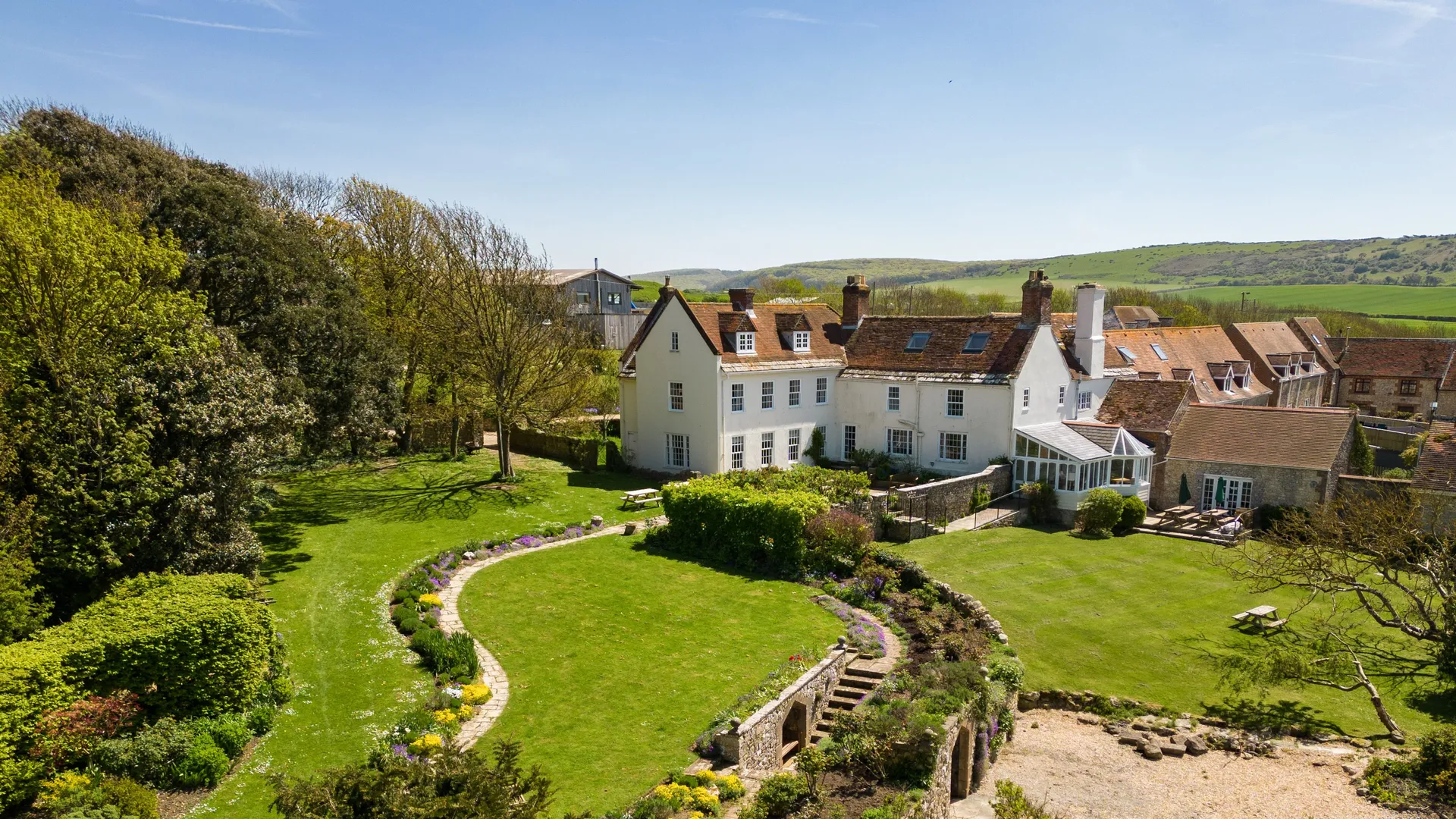Aerial view of Tapnell Manor with manicured gardens, curving flowerbeds and countryside hills under a clear blue sky