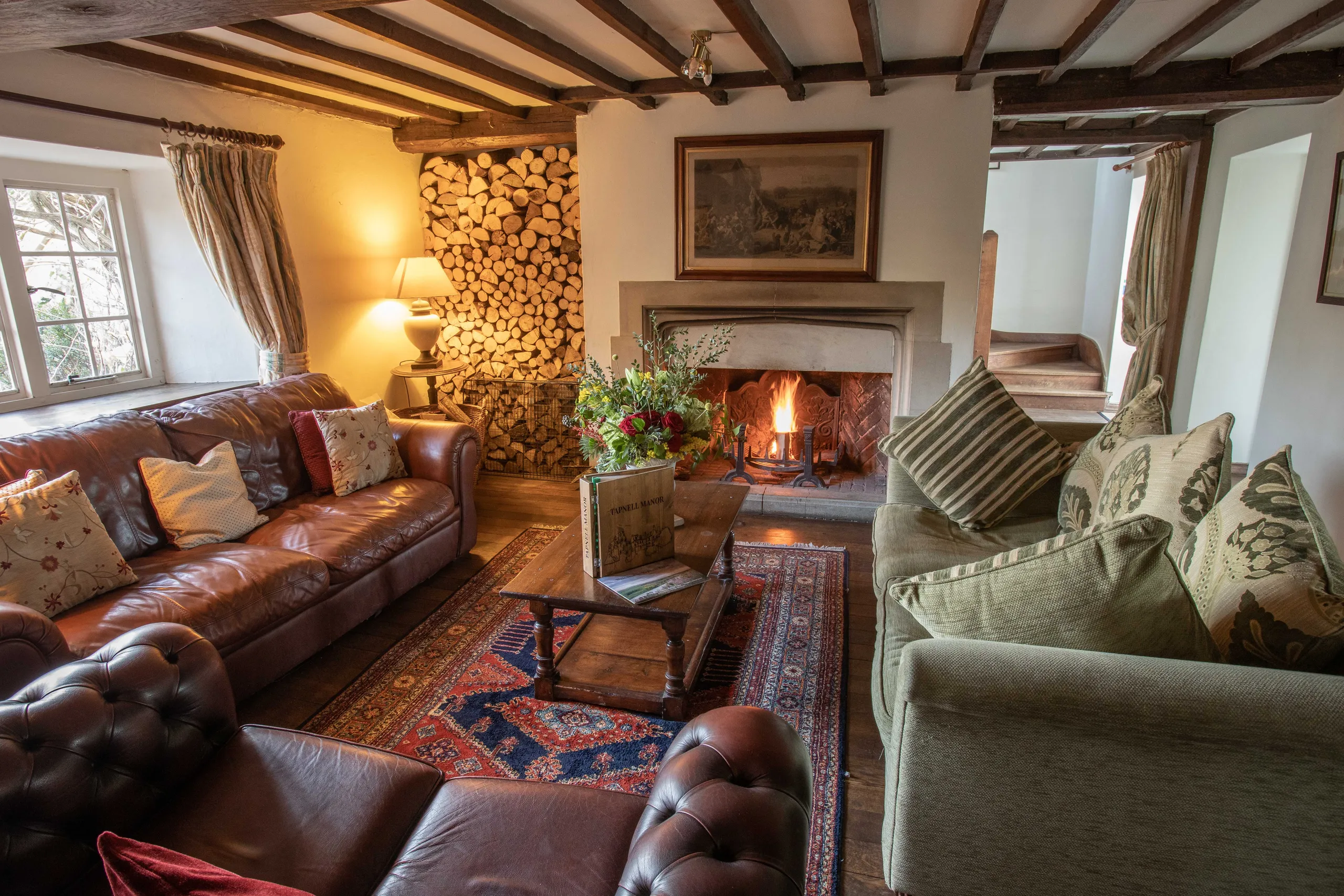 Cosy beamed sitting room with open fire, leather and fabric sofas, and stacked logs at Tapnell Manor
