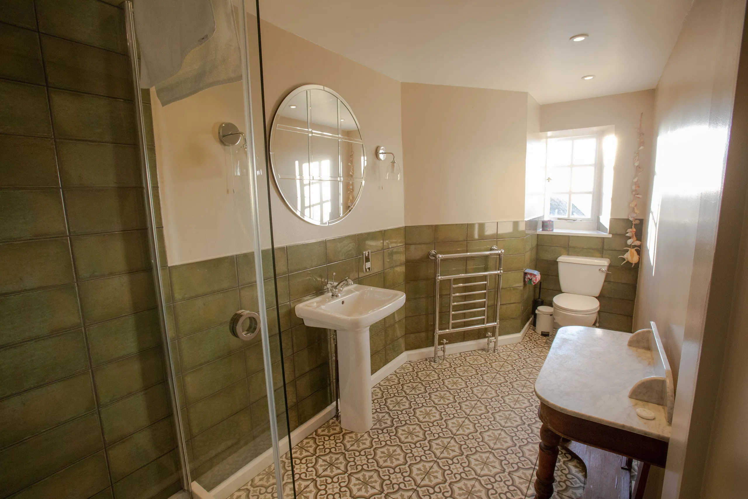 Bright bathroom with green tiled walls, walk-in shower, pedestal sink, round mirror and patterned floor tiles