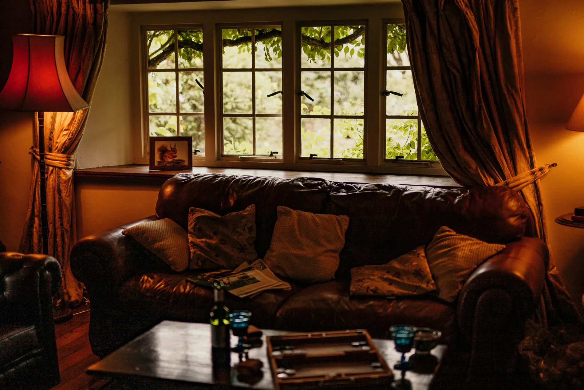Cosy lounge with leather sofa, cushions and table, beside a large window with curtains and leafy view