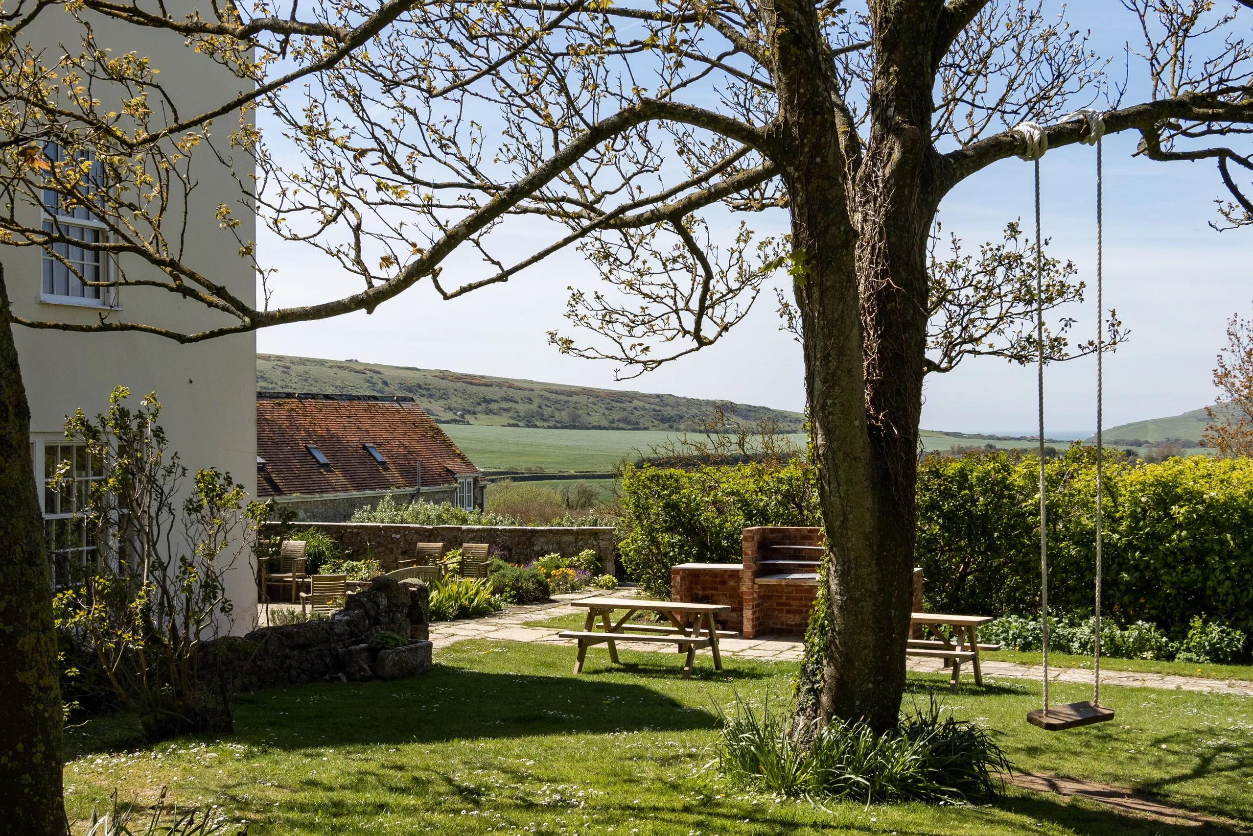 Garden seating area with wooden picnic table and rope swing beneath a tree, overlooking rolling countryside