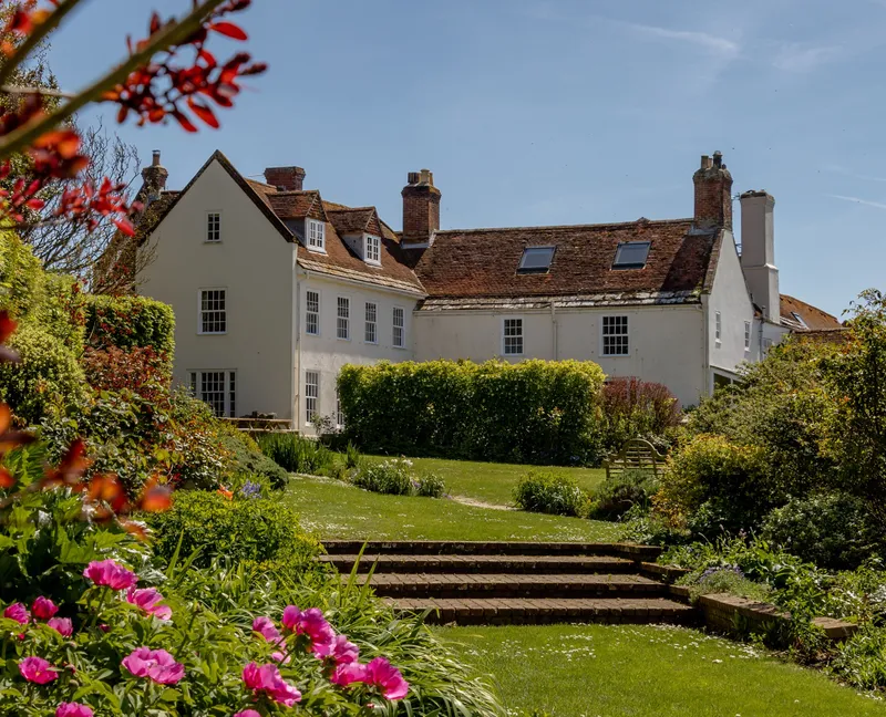 White manor house with red-tiled roof viewed across a sunny garden with steps and pink flowers