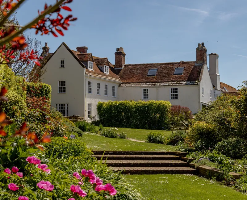 White manor house with red-tiled roof viewed across a sunny garden with steps and pink flowers