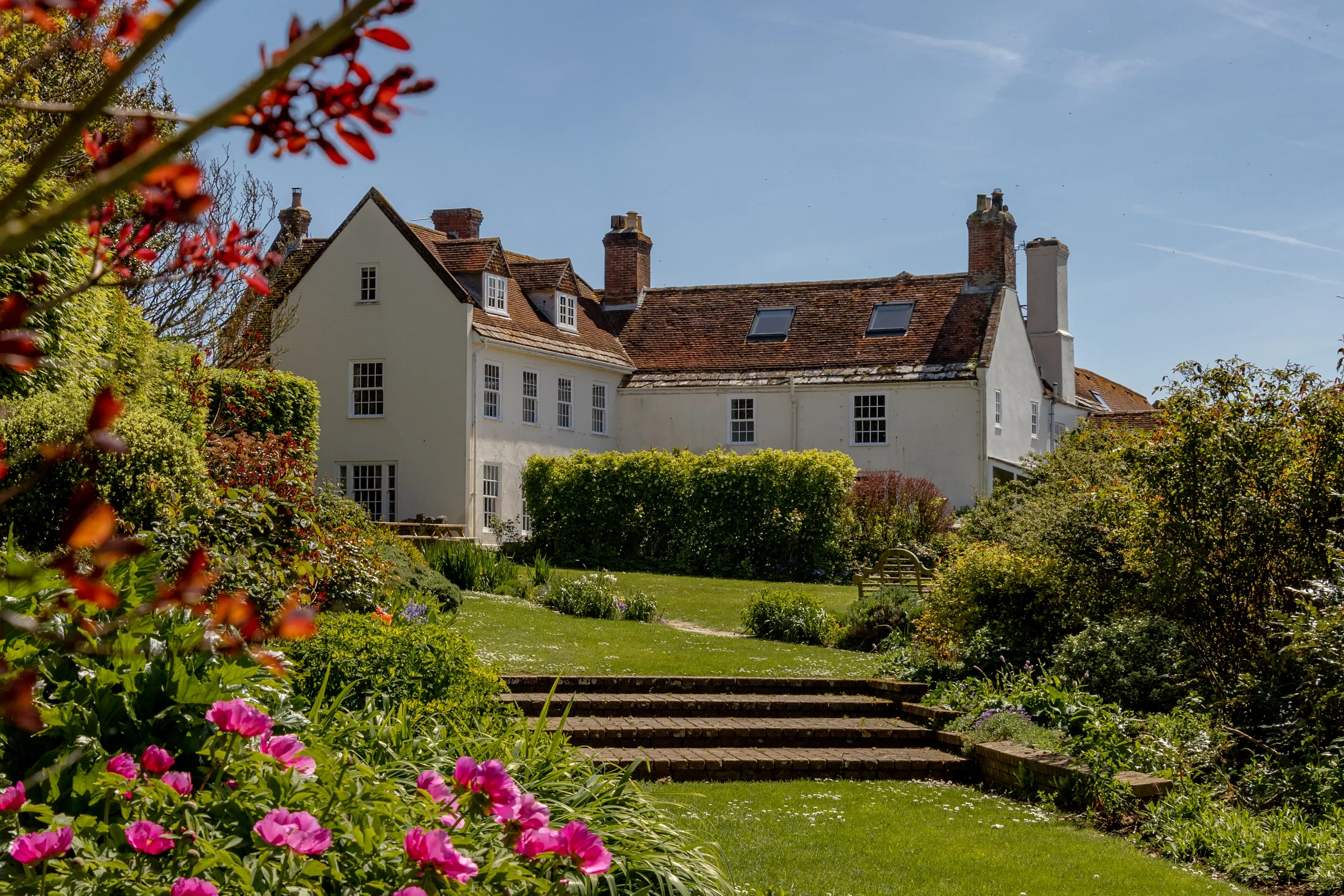 White manor house with red-tiled roof viewed across a sunny garden with steps and pink flowers