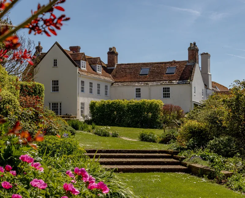 White manor house with red-tiled roof viewed across a sunny garden with steps and pink flowers