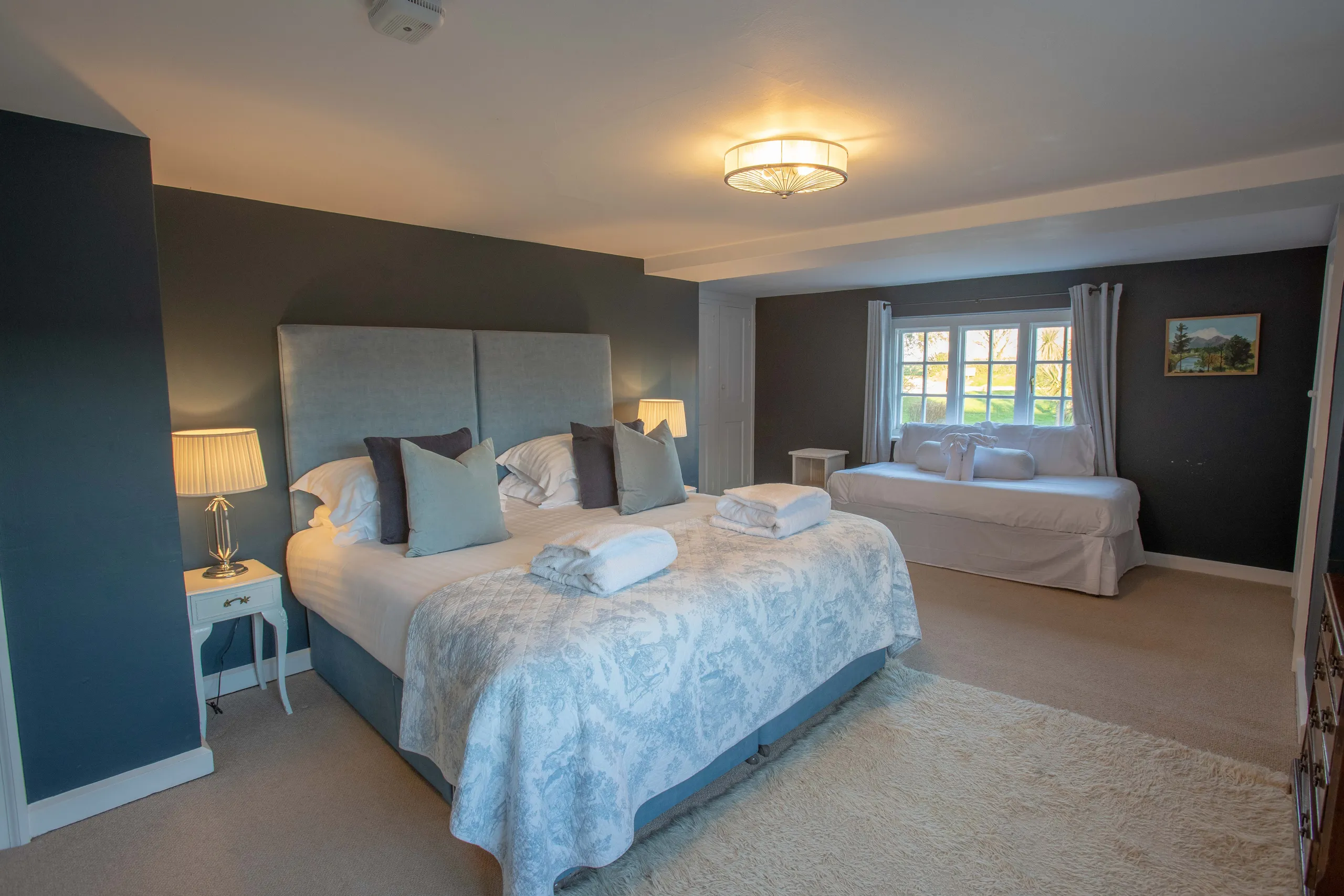 Spacious bedroom with king-size bed, soft blue headboard, bedside lamps and a day bed by the window