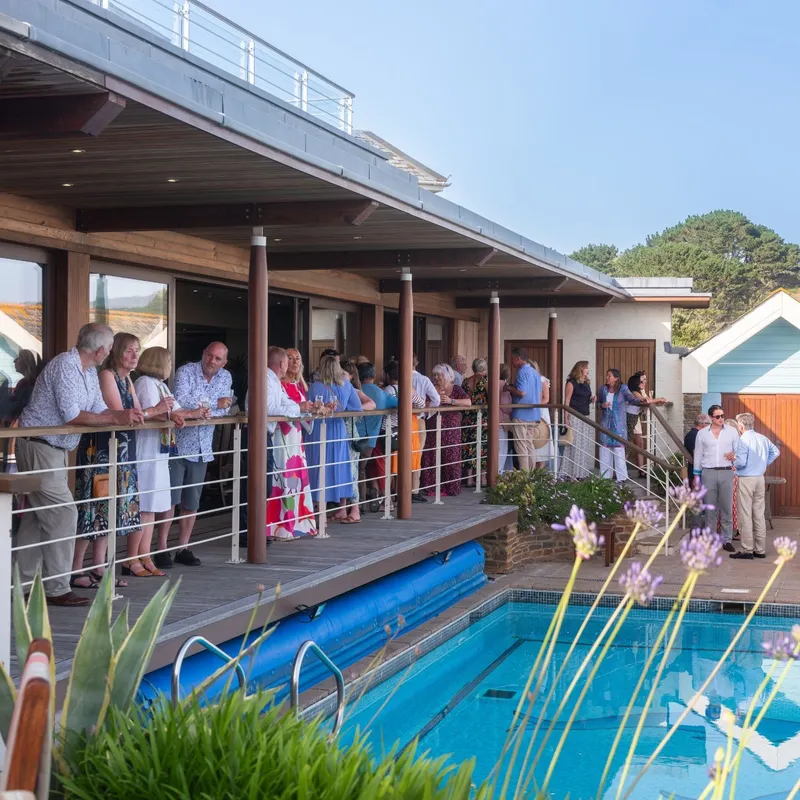 Guests gathered on a wooden terrace beside a swimming pool at a modern countryside home.