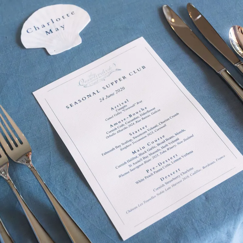 Elegant seasonal supper club menu with silver cutlery and a shell-shaped place card on a blue linen tablecloth.