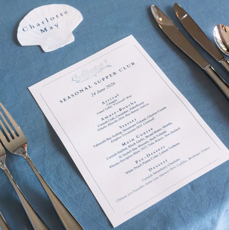 Elegant seasonal supper club menu with silver cutlery and a shell-shaped place card on a blue linen tablecloth.