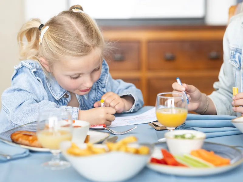 Young girl drawing at a dining table during a meal, with juice, chips and vegetables nearby.