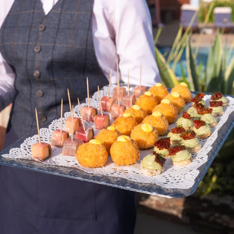 A waiter holding a silver tray of assorted canapés beside an outdoor swimming pool.