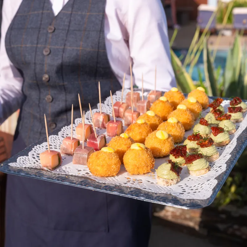 A waiter holding a silver tray of assorted canapés beside an outdoor swimming pool.