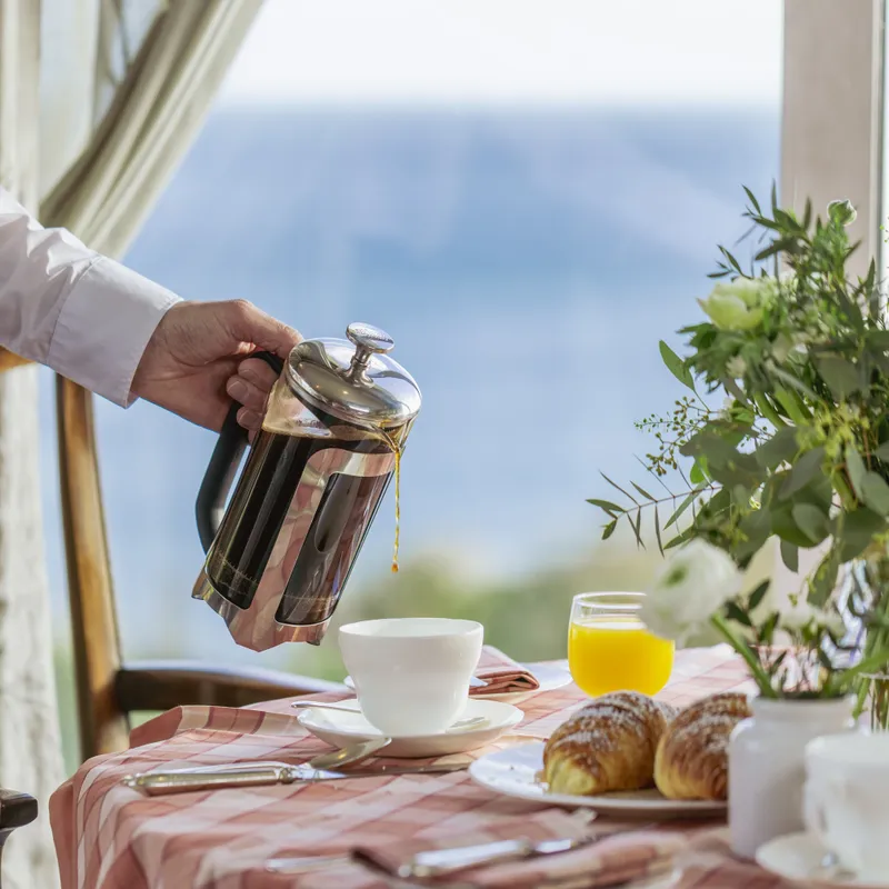 Person pouring coffee from a French press beside a breakfast table with croissants and flowers