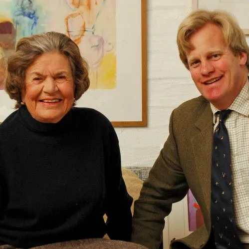 Smiling elderly woman and middle-aged man seated together on a sofa in an art-filled room
