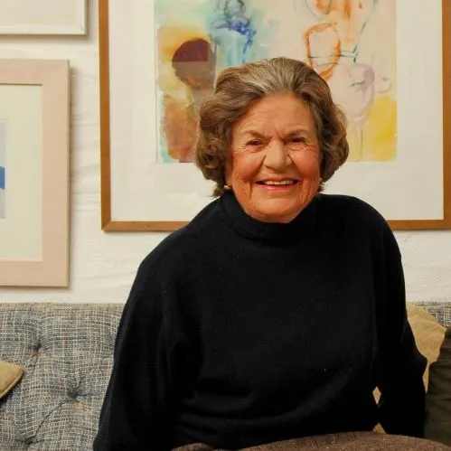 Smiling elderly woman and middle-aged man seated together on a sofa in an art-filled room