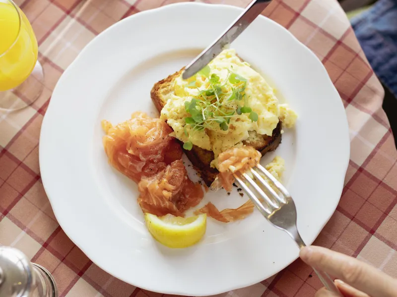 Person cutting into scrambled eggs on toast with smoked salmon and a lemon wedge on a white plate at a café table