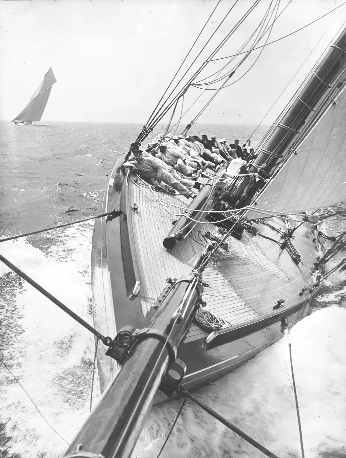 Historic black-and-white photograph of sailors hiking out on the deck of a heeling racing yacht at sea.